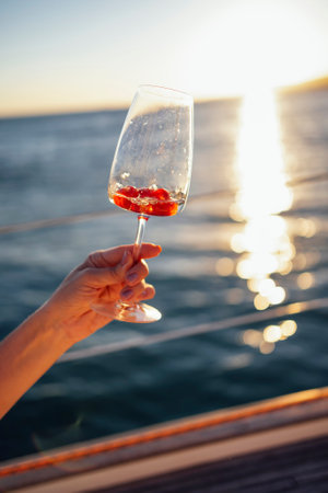 Female hand holding glasses of champagne with raspberries on board the boat. Sunset sky and sea on the background. Making a celebratory toast with sparkling wine. Birthday, holiday, party and friendship conceptの写真素材