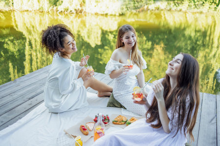 Happy female friends on picnic on the bank of a river. Blanket with pillows and food on a wooden...