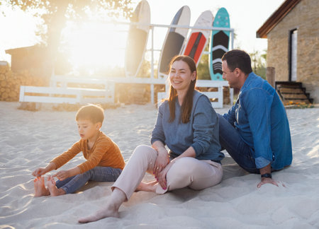 Joyful married couple of surfers with their cute boy on the beach. Smiling man holding paddleboard and hugging woman, holding little son in her arms.の写真素材