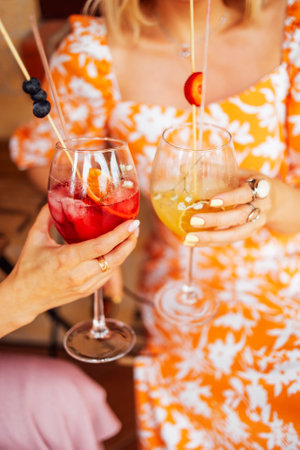 Close up of female hands holding glasses of acohol cocktails with fresh fruts and berries. Making a celebratory toast with mixed drink. Woman in bright orange dress with pattern on the background.の写真素材