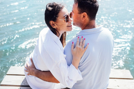 Young couple in love in light casual clothes is sitting on wooden bridge. Happy woman and man hug...