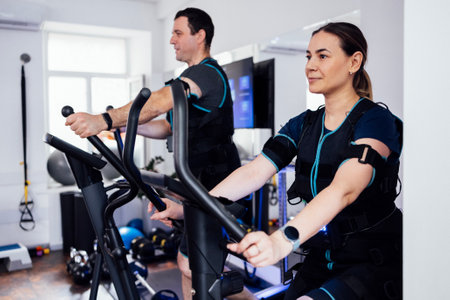 Couple of sports people in ems suits are engaged in sports equipment in fitness club or gym. Laughing young man is exercising on elliptical trainer. Pretty smiling woman pedaling exercise bike.の写真素材