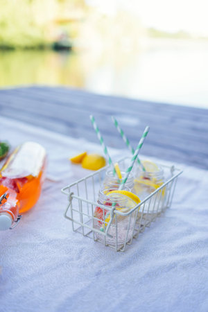 Picnic on the bank of a river. Blanket with pillows and food on a wooden bridge. Bottle of strawberry and lemon smoothie, soda glasses with straws, herb croissants, sweet cakes and fresh strawberriesの写真素材