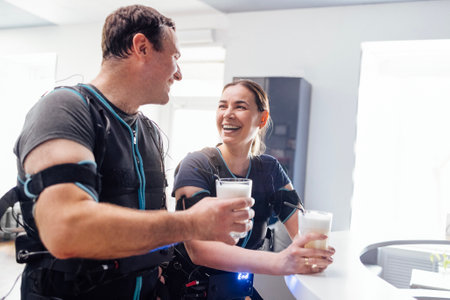 Young smiling man and woman in ems suits communicate and drink protein shakes in sports bar. Rest from training with electrical muscle stimulators. Modern stylish interior of fitness club.の写真素材