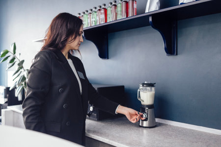 Young pretty woman prepares protein shake using stationary blender. Female barista pours milkshake...