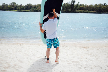 Joyful smiling male surfer on the beach. Young man in light t-shirt and multi colored shorts holding paddleboard. Summertime sport and fitness concept.の写真素材