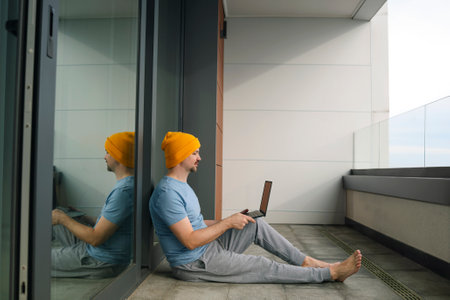 Young man in casual clothes and barefoot sits on balcony with laptop. Male freelancer in yellow hat,...