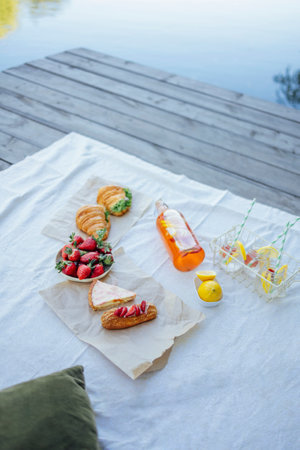 Picnic on the bank of a river. Blanket with pillows and food on a wooden bridge. Bottle of strawberry and lemon smoothie, soda glasses with straws, herb croissants, sweet cakes and fresh strawberriesの写真素材