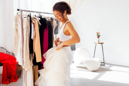 Charming smiling cute african girl in white top and leggings by trempels with stylish clothes. Laughing attractive young afro woman choices of clothing on rack. Shoppingの写真素材