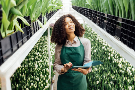 Young smiling mixed race woman uses digital tablet to grow tulips of different types. Female farmer stands on stepladder among natural flowers. Charming gardener works with tulips in greenhouse.の写真素材