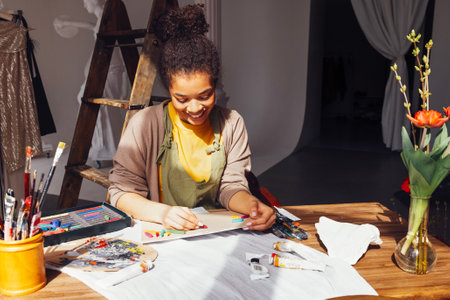 Young creative pretty african american girl artist paints with pastels while sitting at table in...