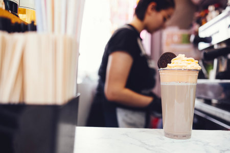 Young brunette female barista in black shirt, apron and black gloves decorates milk cocktail with cream and chocolate cookie. Professional coffee machine and takeaway coffee ingredients on background.の写真素材