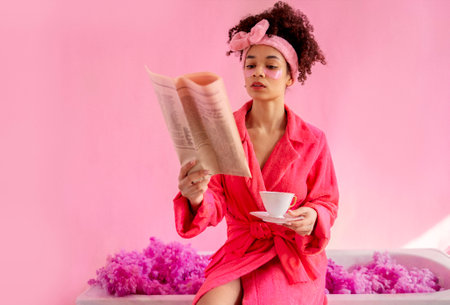 Young attractive female african american with newspaper and porcelain cup. Smiling afro girl with pink patches under her eyes and soft headband reads the news and drinks tea on pink backgroundの写真素材