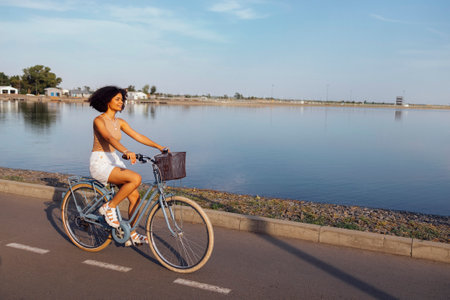 Dark skinned female teenage is driving her bicycle. Young and positive afro american girl in casual cloth smiles sweetly and has a fun. City street and trees with bushes in the background. copy spaceの写真素材