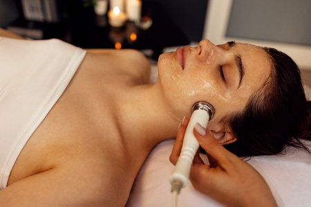 Closeup of face of beautiful relaxing girl undergoing hardware cosmetology procedure in spa. Cream for ultrasonic facial massage is applied to face of young woman using special nozzle in beauty salon.の写真素材