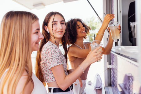 Three cute cheerful teenage girls are buying lemonade in the drinks takeaway store. Happy teens of different nationalities drink cocktails on open air and have fun. summertime. friendship concept.の写真素材