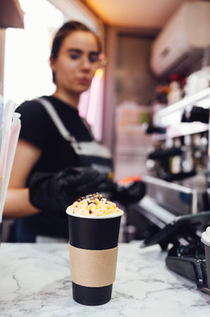 Close up of black paper cup with milk drink with whipped cream and chocolate. Female barista hands in black gloves decorates tasty cocktail with sweet topping. Takeaway coffee preparationの写真素材