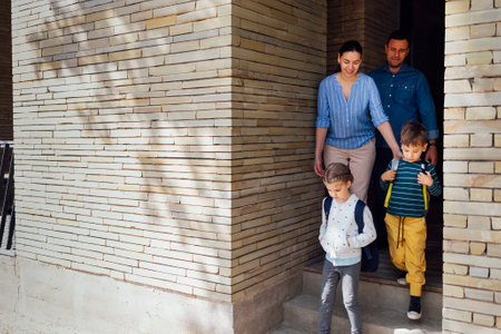 Young parents seeing their cute kids off to school. Smiling mom, dad, daughter and son go down the...