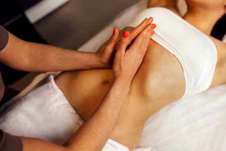 Close up of young woman getting belly massage in spa salon. Female masseuse hands with orange manicure carefully squeeze skin of client. Burning candles on background. copyspace.の写真素材
