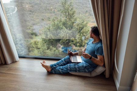 Young man in casual clothes and barefoot sits near panoramic window with laptop. Male freelancer works at computer and drinks tea or coffee. Wonderful view of the green mountains. modern interior.の写真素材
