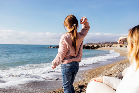 A little girl in a sweater and jeans walks with her family on the seashore. A cute child will throw pebbles into the water. The kid is standing back. Weekends and resting on the beach in autumn.の写真素材