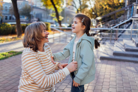 Smiling handsome senior woman in casual clothes and her little cute granddaughter playing on street. Laughing middle aged grandmother and cute little girl having fun outdoors. happy childhood moments.の写真素材