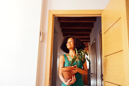 A beautiful girl of african appearance with a pot of a houseplant in her hands walks through the door in a new house. A charming mixed race girl in casual clothes smiles and looks a new apartment.の写真素材