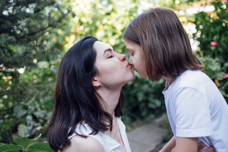 Beautiful brunette woman kissing her little daughter on nose on walk in park. Happy mom and kid is hugging and having a good time together in the garden. Caring mom show tenderness love to her cute kid.の写真素材
