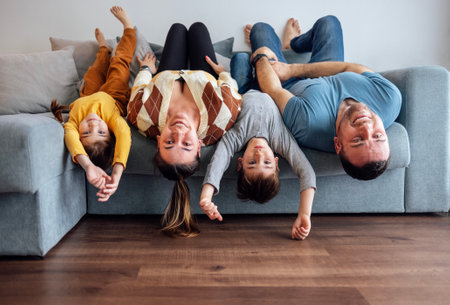 Happy family people and their kids in casual clothes are lying upside down on their backs on gray sofa. Young woman and man, cute boy and girl looking at camera and smiling while playing at home.の写真素材