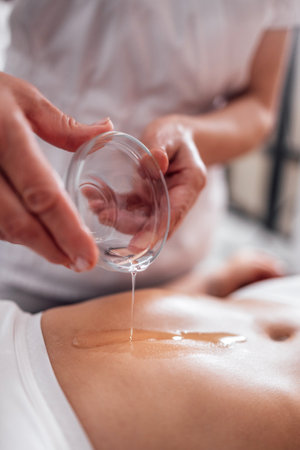 Crop faceless masseuse pouring oil on hand during relaxing massage in spa center. Bodycare, healthcare conceptの写真素材