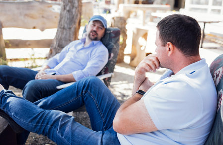 Two young men in casual clothes sit in comfortable chairs outdoors and talking. Handsome people communicate in the cozy backyard of the house. Friends relax and have a good time together. summertime.の写真素材