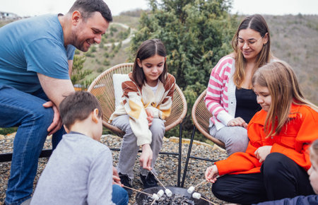 Big happy family roasting mashmallows outdoors. Young mom, dad, three daughters and son have fun in backyard of house and cook delicious treats on fire. Beautiful green hills and trees on background.の写真素材