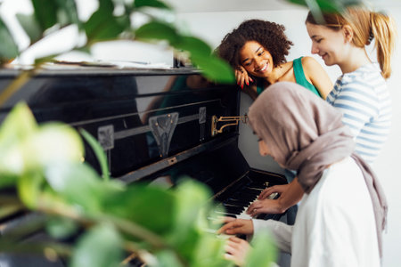 Cute smiling female teenagers play duets on the old piano. Caucasian, afro american and muslim teens are listening to the music. girls have fun. Green leaves on the frontgraund. Free space for text.の写真素材