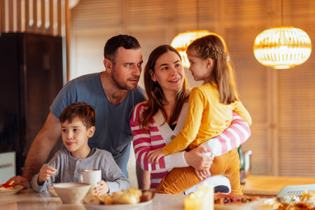 Happy young family has breakfast at the table in the dining room at home. Beautiful mom is holding a sweet cute daughter. Parents are talking to their little child. Funny boy eats and licks his lips.の写真素材