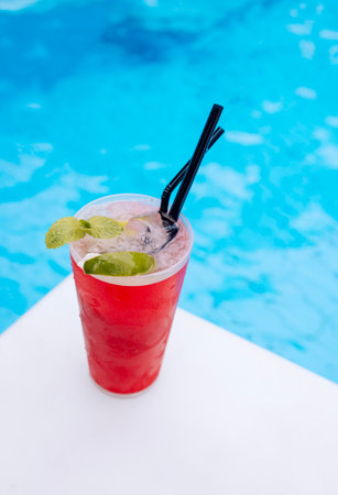 Red glass of refreshing mohito cocktail with two straws near swimming pool during vacation. Cold cup of ice drink with lime slice and mint on background of blue water outdoor. hello summer holiday.の写真素材