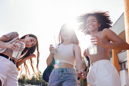 Close up of three teenage girls in casual clothes drinking tasty lemonade on the go. Happy female friends walk outdoors and have fun. Beautiful sunshine and summer timeの写真素材