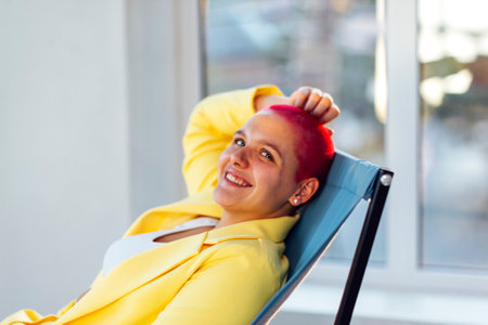 Beautiful relaxed woman in yellow suit and red shot hair holding hand behind head while resting on weekend day at homeの写真素材