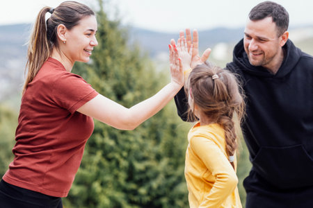 Smiling parents give high five to their little daughter. Happy joyful family in casual clothes have fun in summer park. Man, woman and girl touch each other with their palms.の写真素材