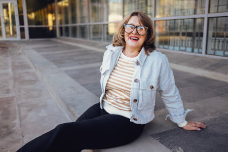 Pretty middle-aged woman in glasses sits on the steps outdoors. Senior woman in casual clothes laughs and looks into the camera. Terrace building with panoramic windows in the background.の写真素材