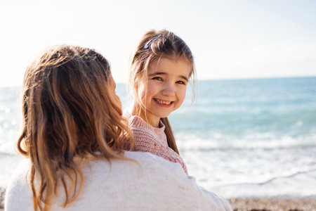 Young mother and little cute daughter in warm sweaters by sea. Blonde woman standing back. Pretty girl smiling and looking at camera. Mom and her baby spend time together. Happy family weekend.の写真素材