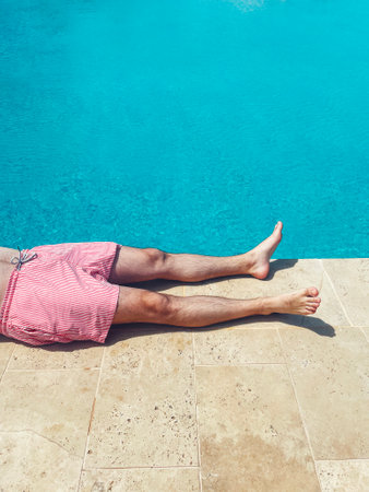 A young man lies on poolside at resort hotel during vacation. Close up of male legs near turquoise water. Sunbathing by the swimming pool. summertime. tourism and travel concept.の写真素材