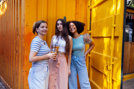 Three cute teenage girls in casual clothes on background of colored shipping container. Multiracial female friends holding cocktails in plastic glasses and smiling. Takeaway shop is in yellow metal boxの写真素材