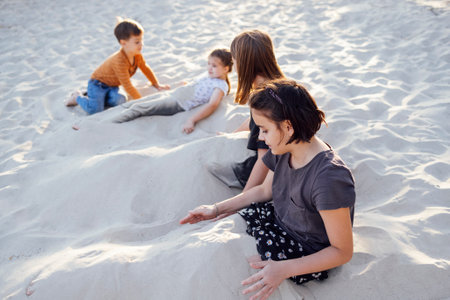 Four joyful children in casual clothes play on the beach. Funny girls pour sand on themselves. Smiling sisters dig their legs into sand. Cute kids have fun near the sea. Family vacation and happy childhood.の写真素材
