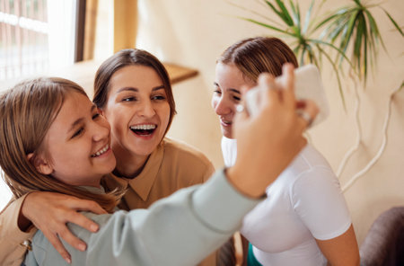Three cute smiling teen girls hugging and taking a selfie. Female teenagers laughs and takes a photo of themself on the phone. Cheerful girls in casual clothes have fun at home.の写真素材