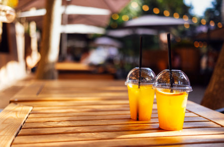 Two orange color cocktails in plastic glasses with straws against the backdrop of an open air cafe. Cold lemonades with lemon slices on a wooden table. Cafe interior on the background. copyspace.の写真素材