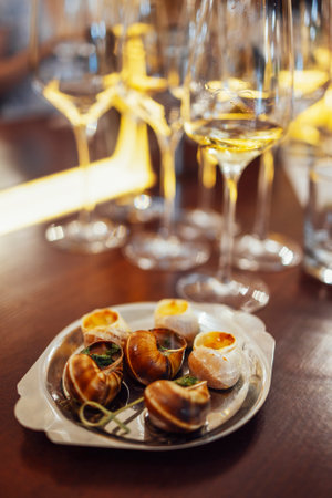 An exquisite delicacy of snails on a metal tray. Escargot with herbs on a platter on a wooden table. Delicious bourguignonne snail. Wine glasses on background. gastronomic tourism. Health nutrition.の写真素材