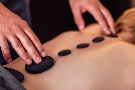 Close-up of the male hands of a masseur doing a massage with hot stones to his client woman. A large black pebble on the female back, along the spine. The concept of relaxation and selfcare.の写真素材