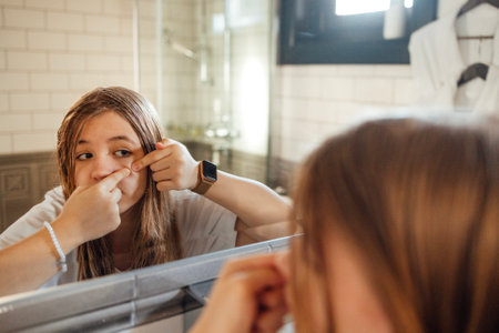 Cute teenage girl squeezes out a pimple on her cheek in the bathroom. A teen in casual clothes with problematic skin touches her face. reflection in the mirror. Upset teenager. copyspace.の写真素材
