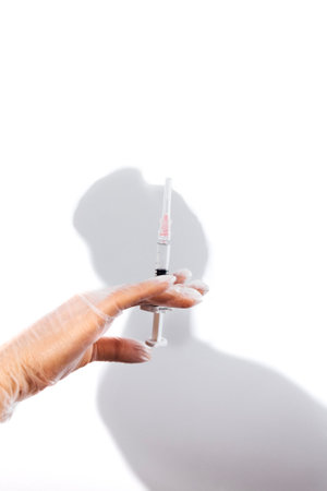Close up of woman hand in medical transparent gloves holding syringe with clear liquid. White background with female shadow. Medical treatment, vaccination, filler injection, cosmetic surgical conceptの写真素材