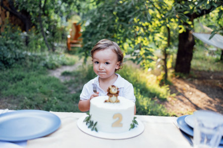 Cute toddler boy with wavy blond hair and white shirt sits at table near birthday cake outdoors. Festive pie with lion cub and number two. Little child tastes holiday sweetness in the garden at home.の写真素材